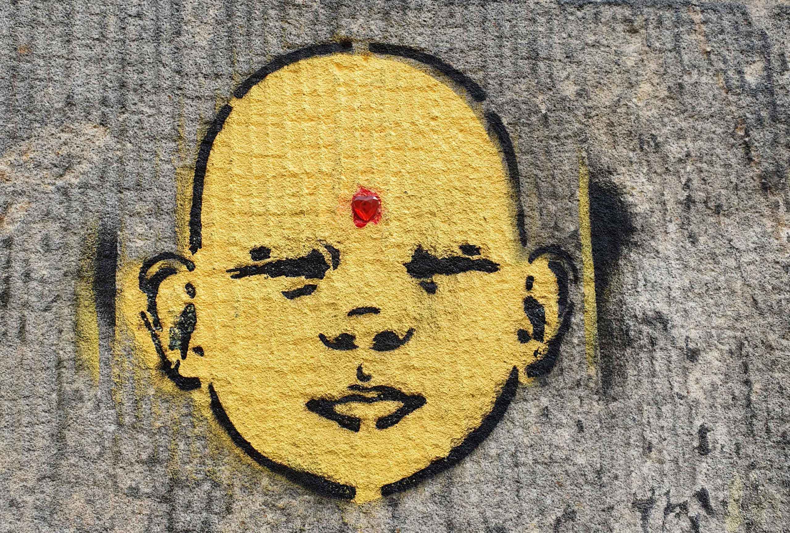 Street Art: gemaltes gelbes Kindergesicht mit rotem Herz-Bindi auf der Stirn